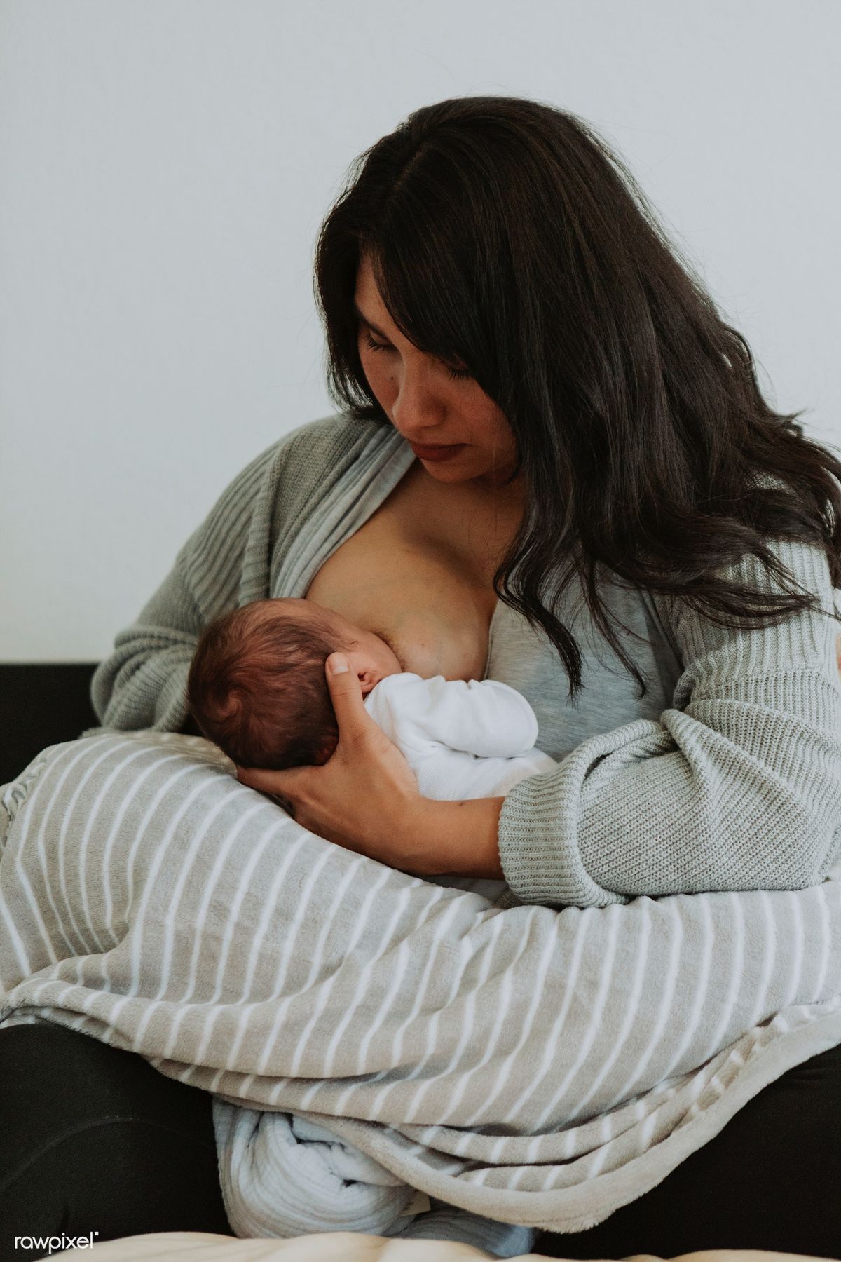 A woman with long dark hair breastfeeding a newborn, who rests on a striped grey pillow across her lap.