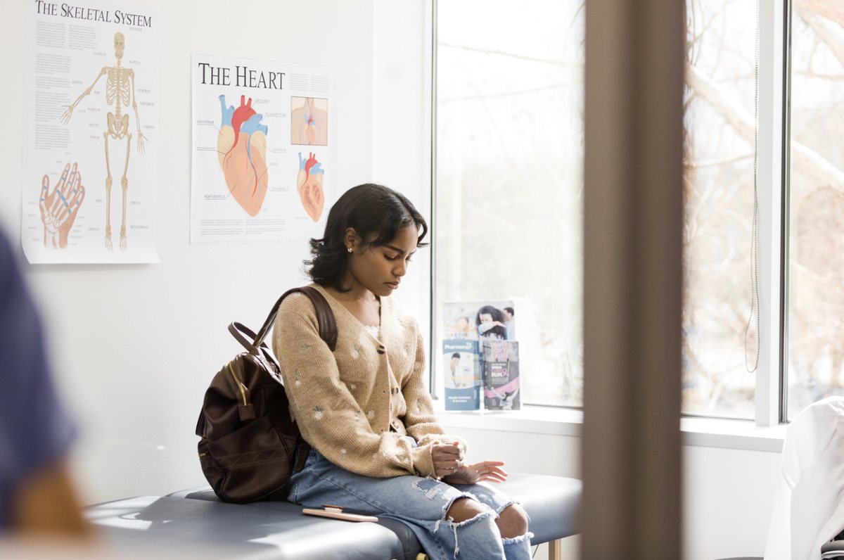 A young woman with a backpack sitting on an examination table in a clinic room, with anatomy posters of the skeleton and the heart on the wall behind her.