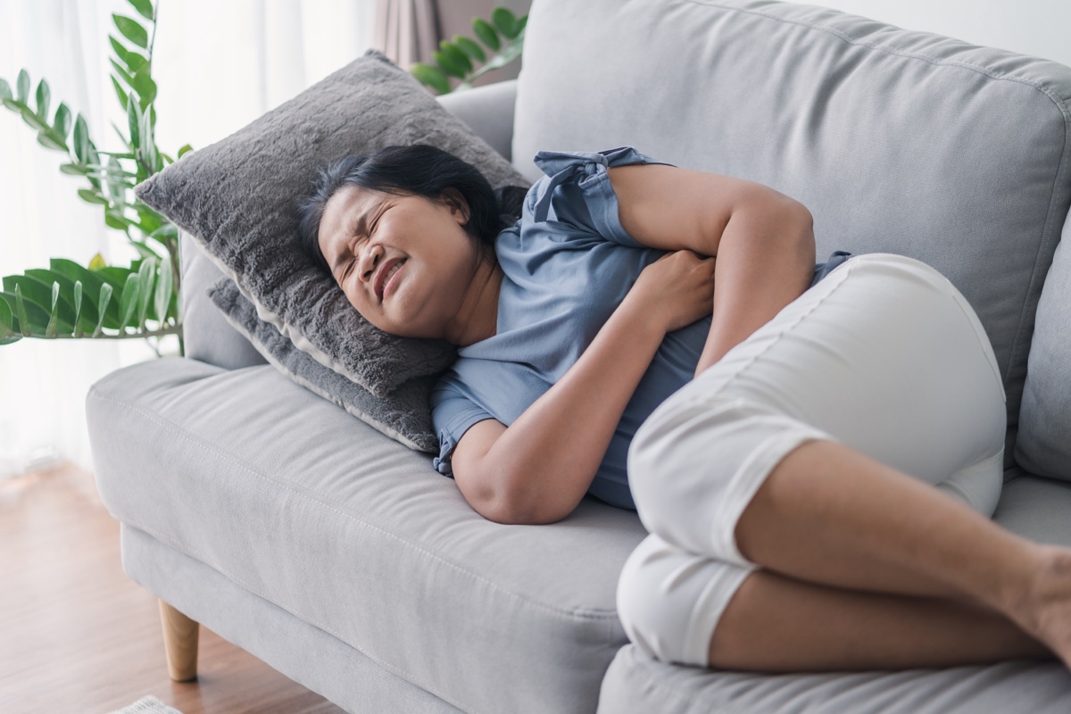 A woman lying on her side on a grey sofa with her eyes closed, holding her abdomen with both arms.