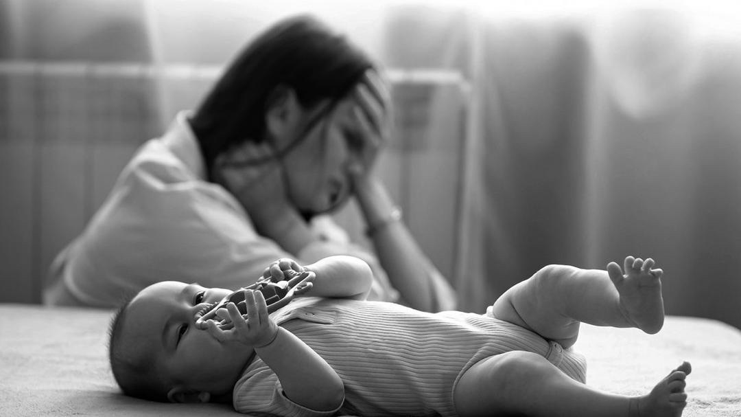 Black-and-white photograph of a baby lying on a bed in the foreground, with a woman seated behind, one hand raised to her head.